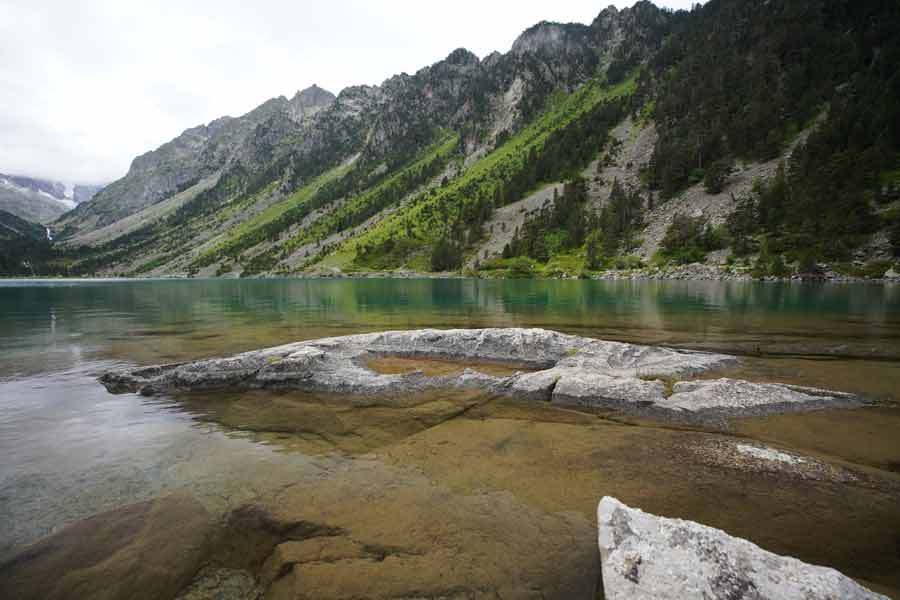 Rives calmes du lac de Gaube par temps couvert, avec eau transparente, rochers immergés et pentes verdoyantes des Pyrénées
