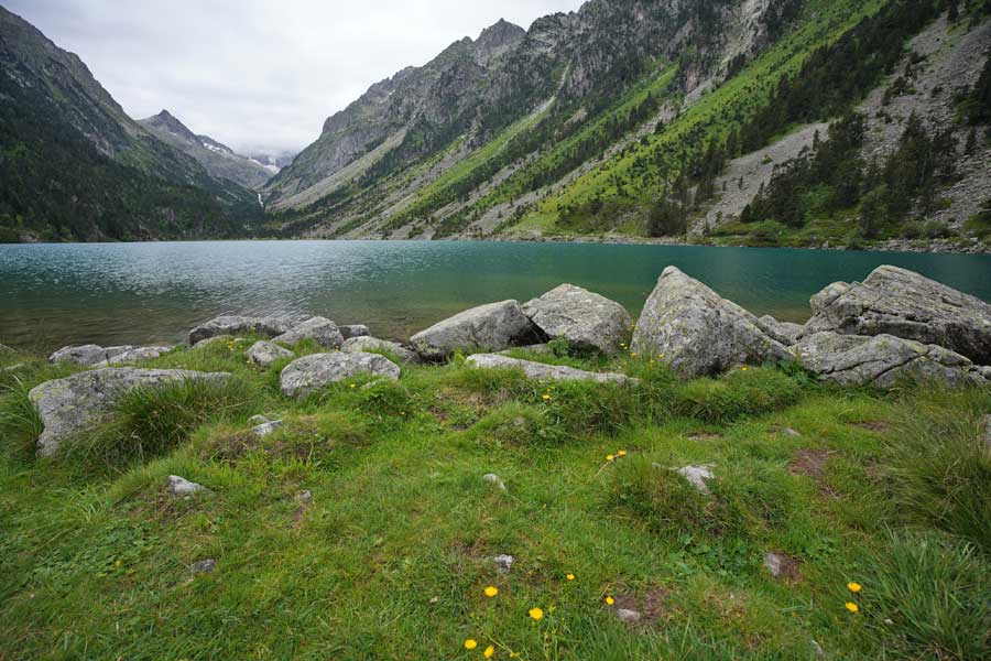 Vue d’ensemble du lac de Gaube depuis la rive, avec eau turquoise, rochers, prairie alpine et montagnes des Pyrénées par temps nuageux