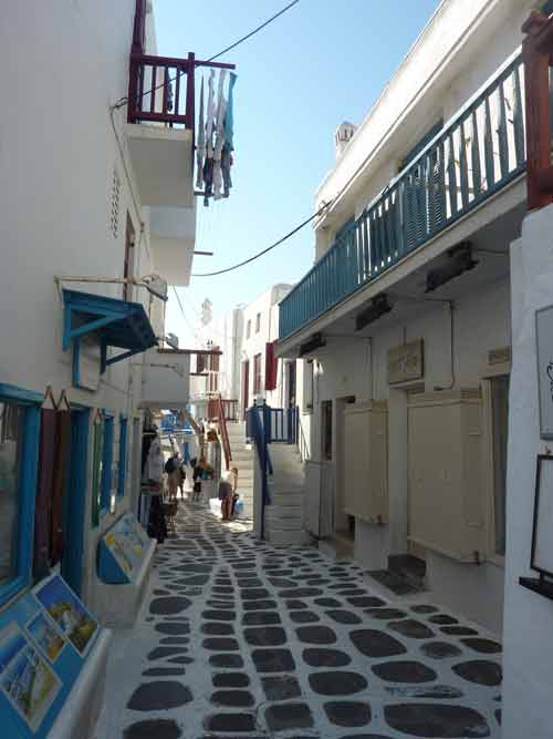 Ruelle étroite et pavée de Chora à Mykonos, bordée de maisons blanches aux volets bleus et de petites boutiques