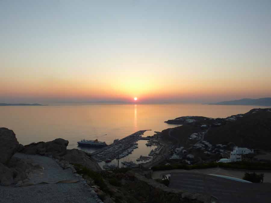Coucher de soleil spectaculaire sur la mer Égée vu depuis le "sunset living room" du Manolia View à Mykonos, avec le nouveau port et ses ferries en contrebas baignés dans une lumière dorée