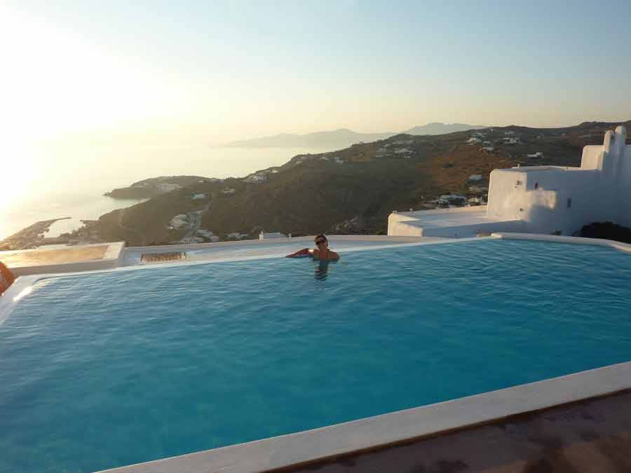 La piscine à débordement du Manolia View à Mykonos avec sa vue panoramique sur la mer Égée et les collines environnantes au coucher du soleil