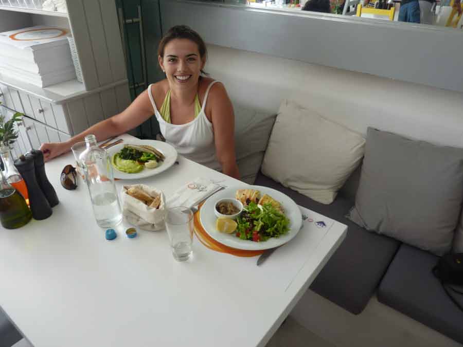 Une voyageuse souriante attablée devant un repas grec généreux au restaurant Avli tou Thodori à Platis Gialos, Mykonos, avec mezze et salades fraîches servis sur fond de décor blanc épuré