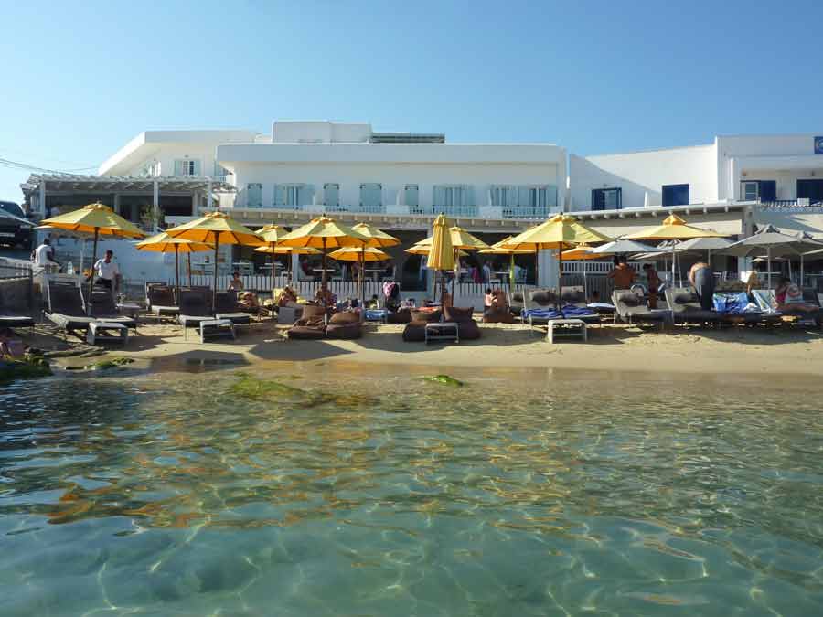 La plage privée du restaurant Avli tou Thodori à Platis Gialos, avec ses transats et parasols jaunes alignés au bord de l'eau turquoise, devant la façade blanche de l'établissement