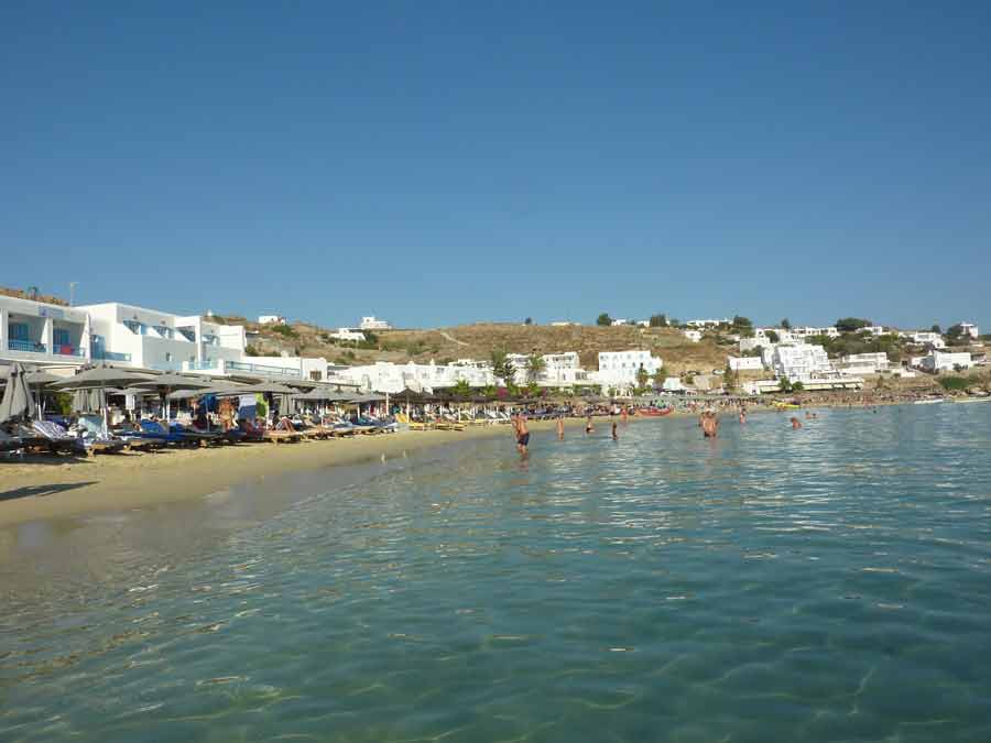 Vue depuis l'eau de la plage de Platis Gialos à Mykonos, avec ses transats colorés, ses baigneurs et ses constructions blanches cycladiques sur la colline en arrière-plan