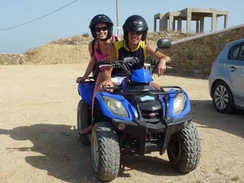 Deux voyageurs casqués sur un quad bleu pour visiter Mykonos, avec les ruines de Délos visibles en arrière-plan