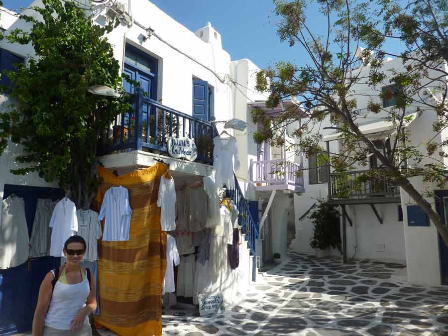 Ruelle pavée de Chora à faire à Mykonos, bordée de maisons blanches aux volets bleus et lilas, avec des boutiques de vêtements en plein air et leurs étoffes colorées suspendées à l'entrée