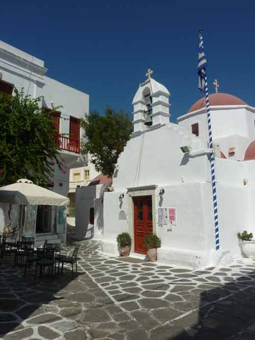 Petite église orthodoxe blanche à dôme rose sur une placette pavée de Chora à visiter Mykonos, avec un clocher orné du drapeau grec