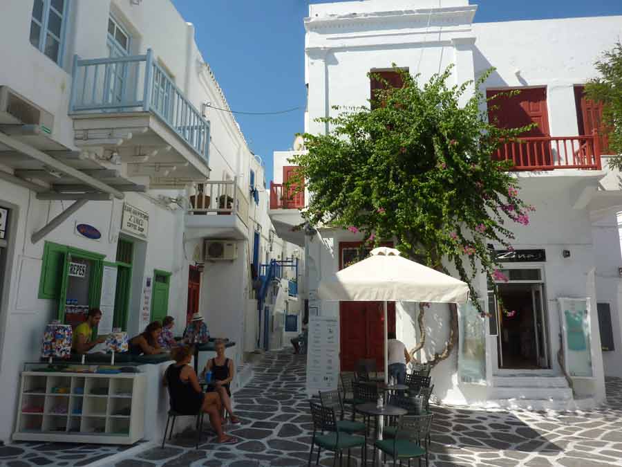 Petite place animée de Chora à Mykonos avec ses maisons blanches aux volets colorés, une terrasse de café ombragée et des touristes attablés sous un arbre en plein soleil
