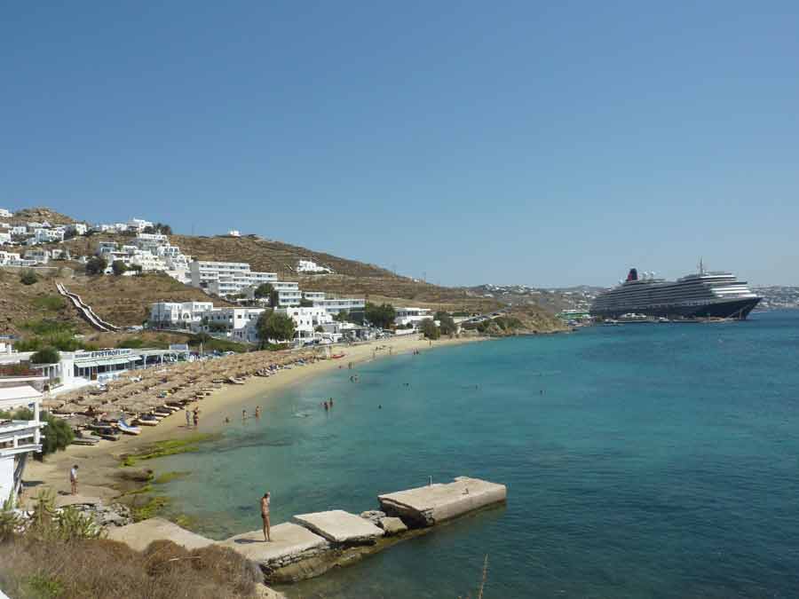 Vue plongeante sur la plage d'Agios Stefanos à Mykonos, avec ses transats sous parasols en paille, ses eaux turquoise et un grand paquebot de croisière ancré dans la baie