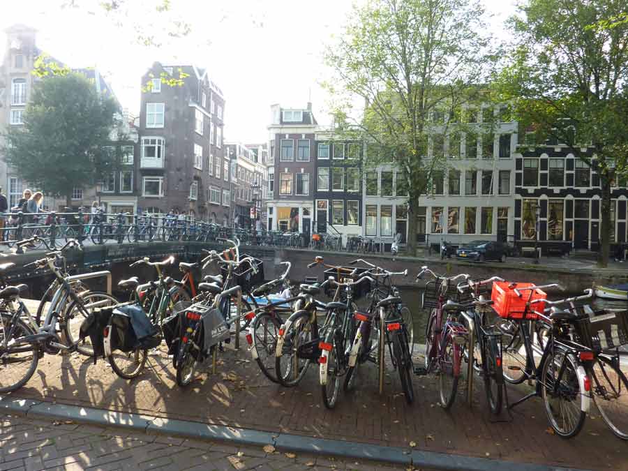 Vélos stationnés le long d’un canal pour visiter Amsterdam avec pont et maisons typiques en arrière-plan