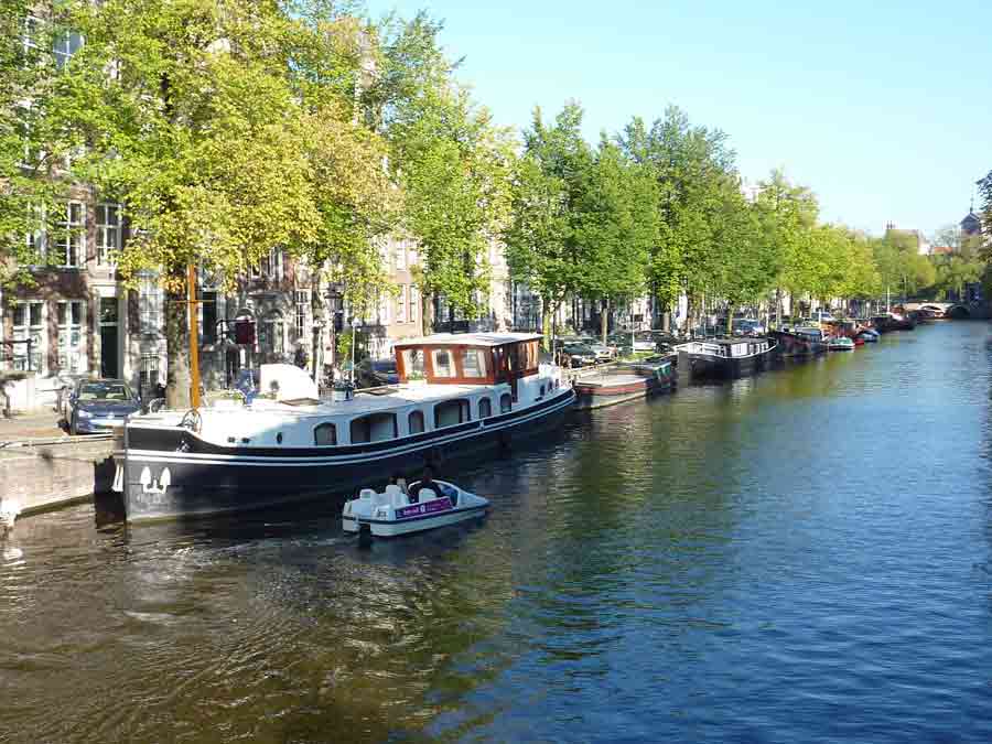Bateaux amarrés sur un canal d’Amsterdam bordé d’arbres et de maisons typiques
