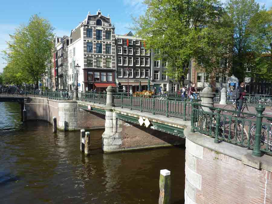 Pont en fer sur un canal d’Amsterdam avec bâtiments anciens et passants en arrière-plan qui visitent amsterdam
