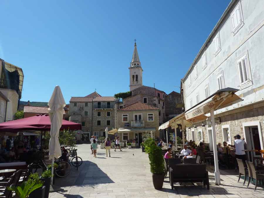 La place centrale de Stari Grad avec ses terrasses animées, ses façades en pierre dorée et le clocher de l'église sous un ciel bleu éclatant