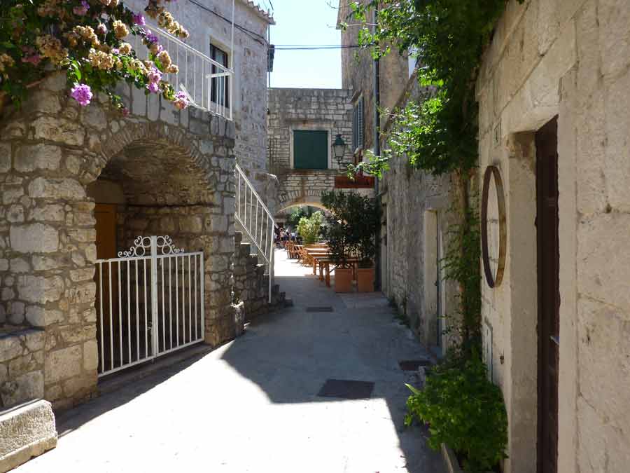 Ruelle pavée de Stari Grad bordée de maisons en pierre blanche et de bougainvilliers, avec une terrasse de restaurant au fond, Hvar, Croatie