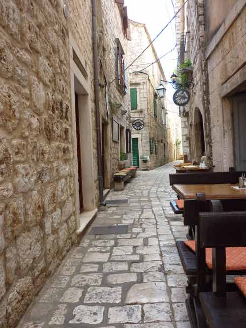 Ruelle médiévale pavée de Stari Grad bordée de tables de restaurant et de façades en pierre blanche, Hvar