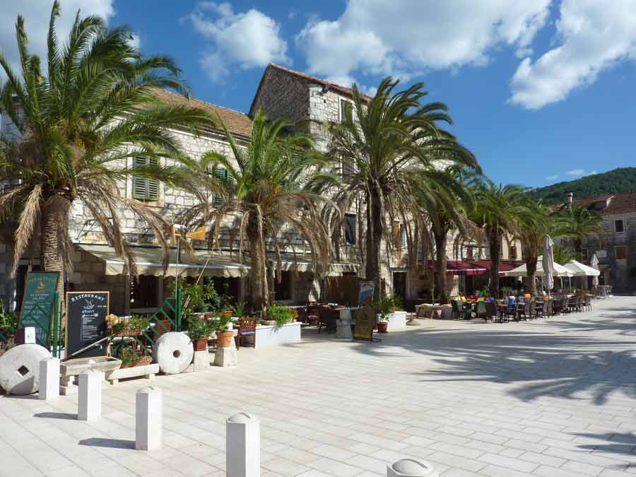 Place principale de Stari Grad bordée de palmiers et de restaurants en terrasse, façades en pierre dorée sous un ciel bleu d'été, Hvar, Croatie