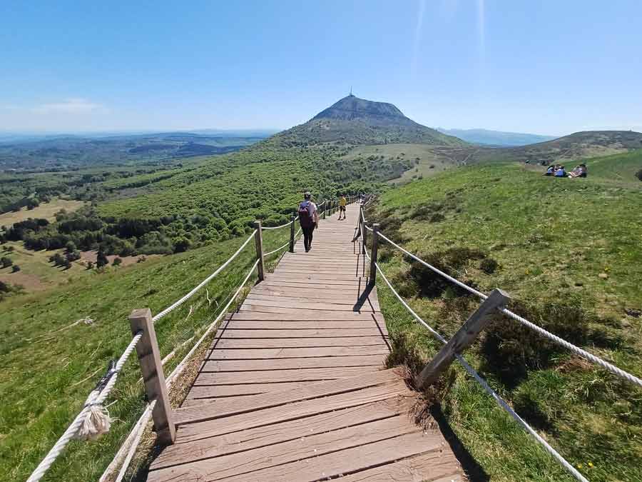 Escaliers en bois aménagés sur le versant du puy Pariou avec vue panoramique sur le puy de Dôme et ses antennes au sommet, randonneurs sur le sentier balisé de la Chaîne des Puys en Auvergne par ciel bleu dégagé