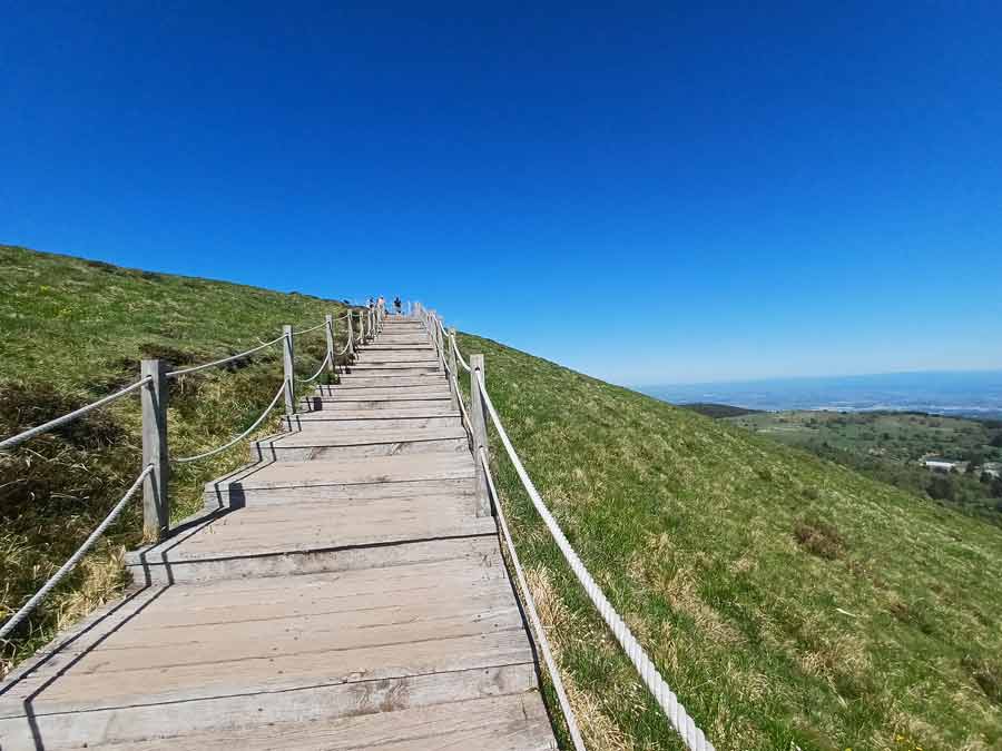 Escaliers en bois avec rampes de corde longeant le flanc herbeux du puy Pariou sous un ciel bleu azur, sentier aménagé pour préserver les sols volcaniques de la Chaîne des Puys en Auvergne