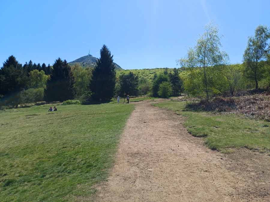 Sentier de randonnée en terre au départ du parking des Goules avec vue sur le puy de Dôme et ses antennes en arrière-plan, large chemin balisé accessible aux familles dans la Chaîne des Puys en Auvergne