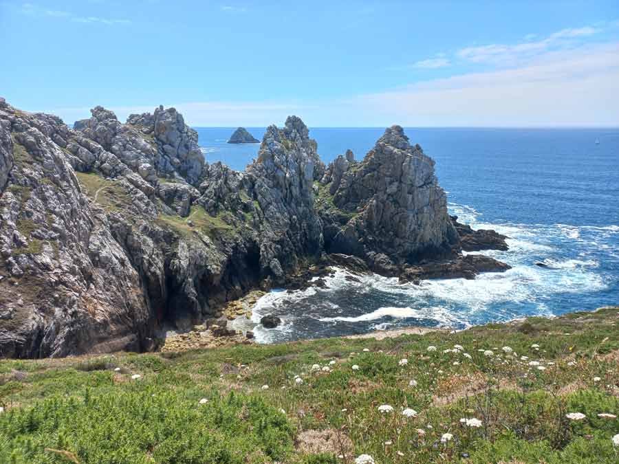 Crique sauvage encadrée de falaises rocheuses à la pointe de Pen Hir, avec les Tas de Pois en arrière-plan et une végétation de lande fleurie au premier plan