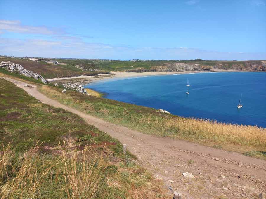 Chemin de randonnée surplombant une crique aux eaux turquoise avec deux voiliers au mouillage, aux abords de Camaret-sur-Mer, presqu'île de Crozon, Bretagne