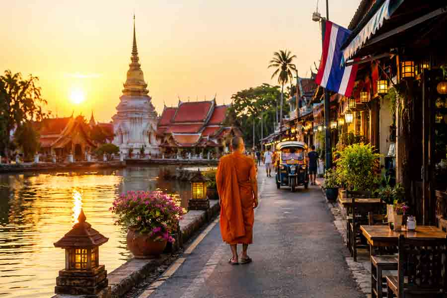 Moine bouddhiste marchant le long d'un canal au coucher du soleil devant un temple thaïlandais, avec tuk-tuk et drapeaux thaïlandais en arrière-plan